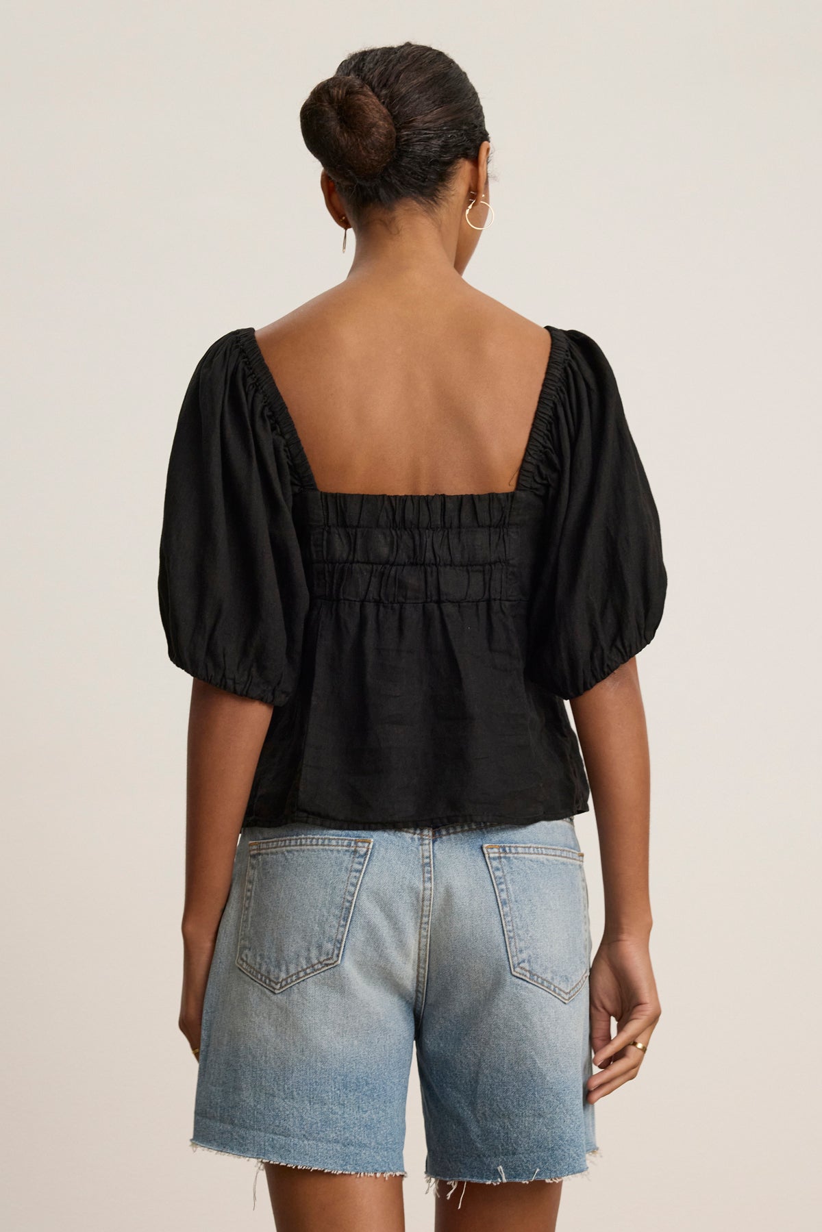 A person with their hair in a bun models the OLIVIA LINEN TOP by Velvet by Graham & Spencer, featuring black puff sleeves and a square neckline, paired with denim shorts and shown from the back against a plain background.