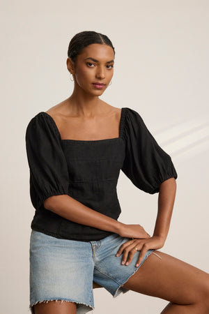 Someone wearing the OLIVIA LINEN TOP by Velvet by Graham & Spencer, a black woven linen piece with puff sleeves and a square neckline, sits on a stool in light denim shorts against a plain backdrop.