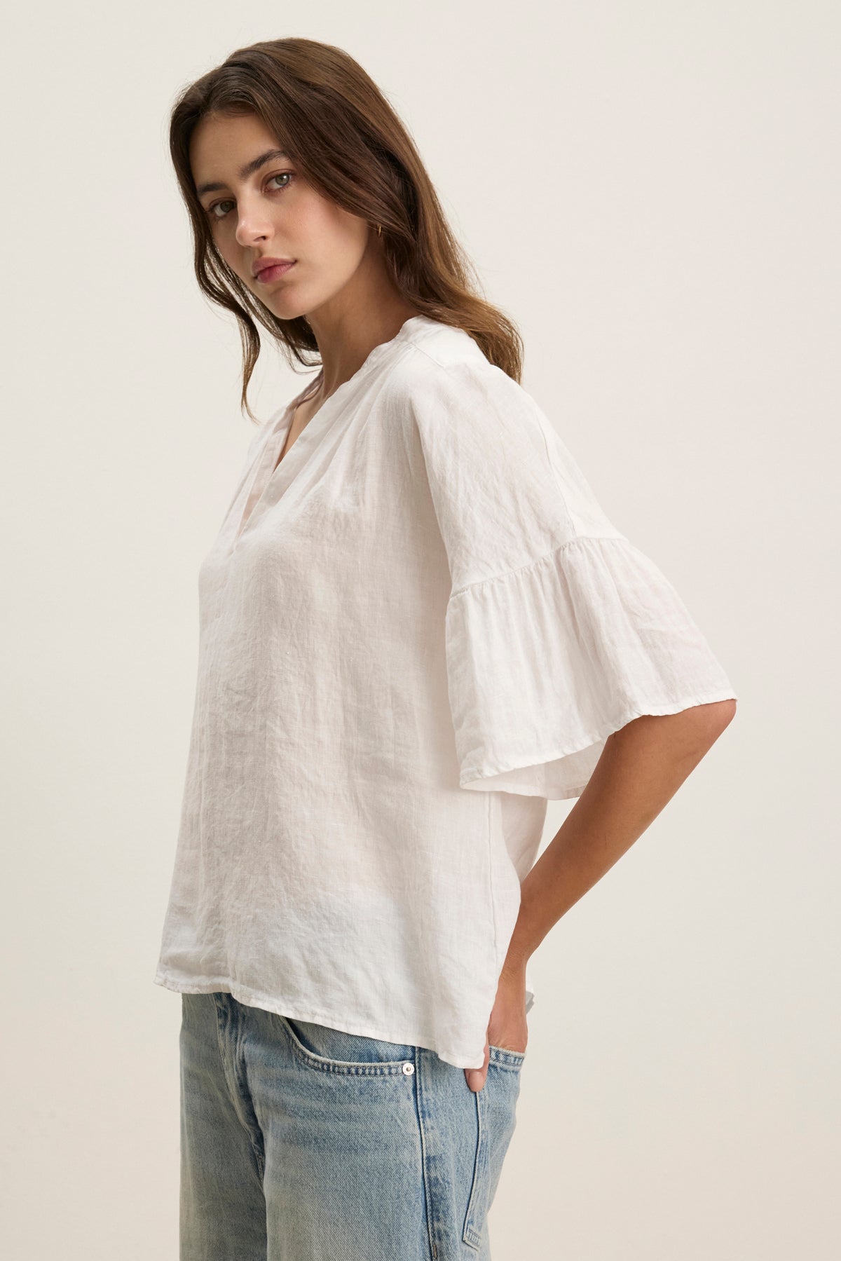 Woman wears the RAYE LINEN TOP by Velvet by Graham & Spencer, a loose woven linen blouse with flared sleeves and an oversized fit, paired with light blue jeans against a plain light background.-39111855538369
