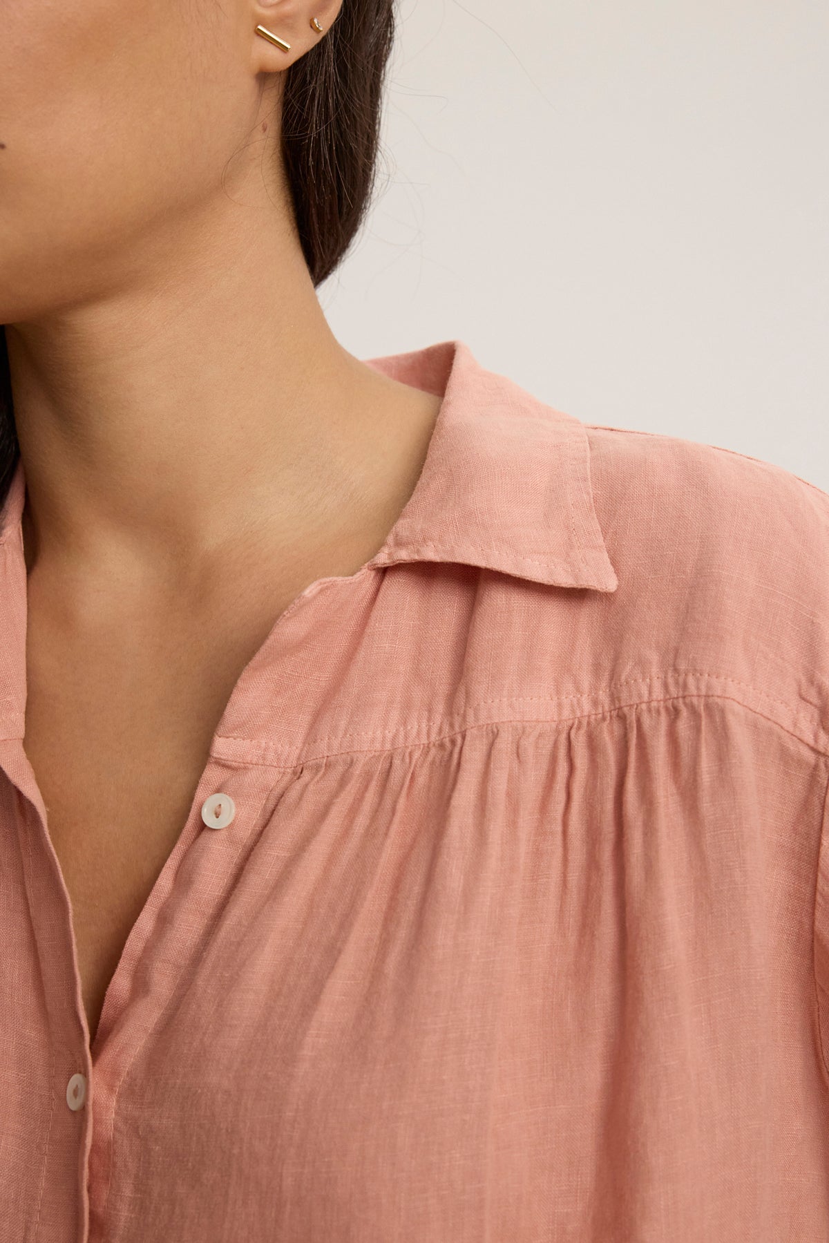A woman wears the ROWAN LINEN TOP by Velvet by Graham & Spencer—a light pink, cropped, button-front collared blouse—paired with a gold ear cuff and shown from the shoulder up against a neutral background.-39123405078721