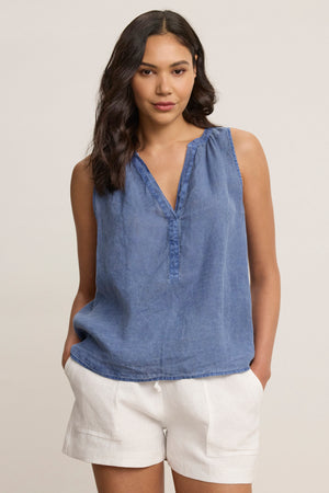 A woman wears the TACY LINEN TANK TOP by Velvet by Graham & Spencer, a sleeveless blue woven blouse with a v-neckline, paired with white shorts, standing with her hands in her pockets against a plain background.