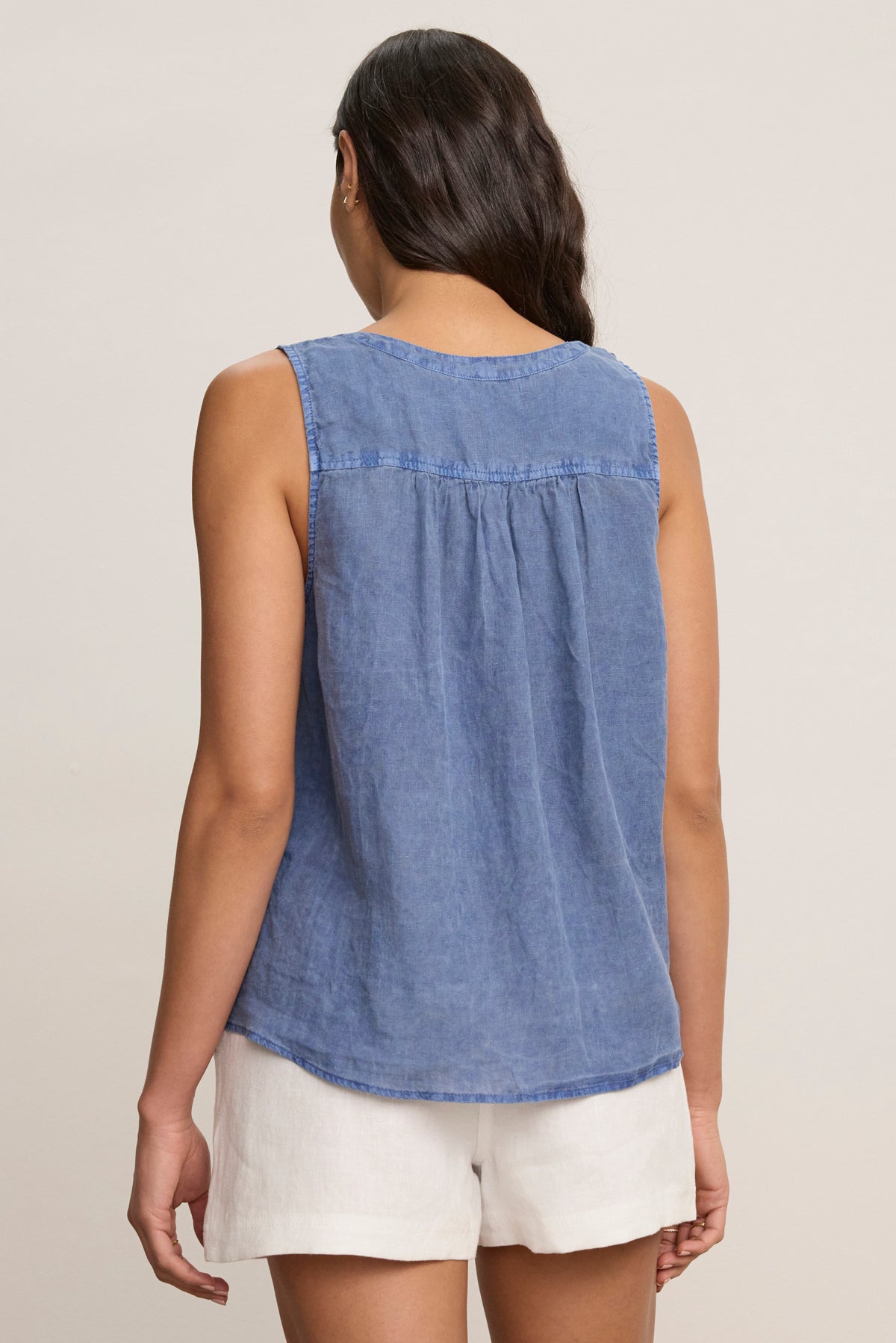 A person with long dark hair wears the TACY LINEN TANK TOP by Velvet by Graham & Spencer, a sleeveless blue woven linen top, paired with white shorts and stands with their back to the camera against a plain light background.
