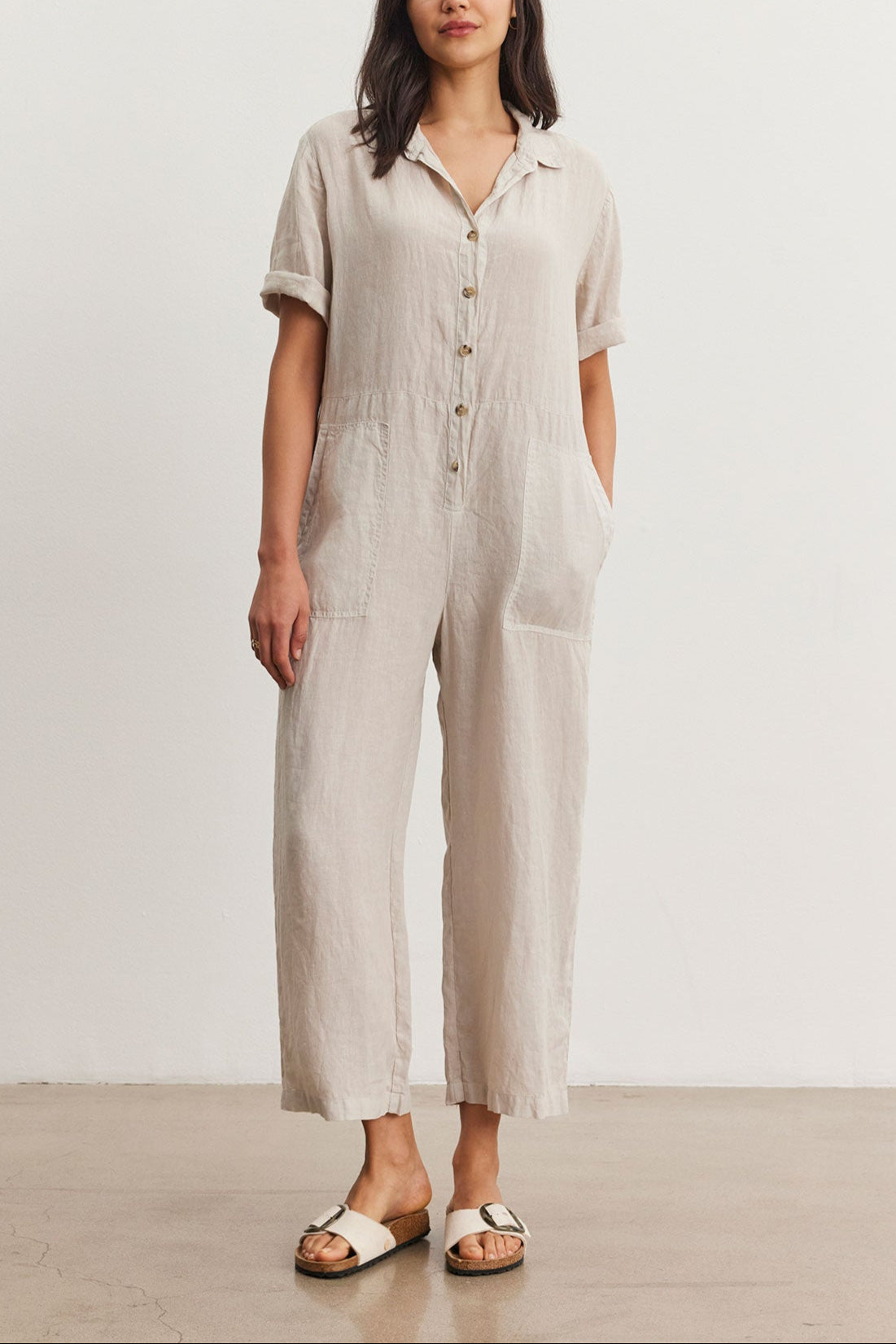 A woman models a casual beige Velvet by Graham & Spencer DELILAH LINEN JUMPSUIT with short sleeves and front buttons, paired with white sandals, standing in a simple studio setting.-36998726549697