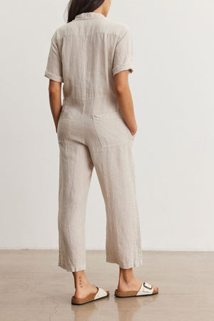 Woman standing facing away, wearing a beige short-sleeved Velvet by Graham & Spencer DELILAH LINEN JUMPSUIT and white sandals, in a minimalist room.
