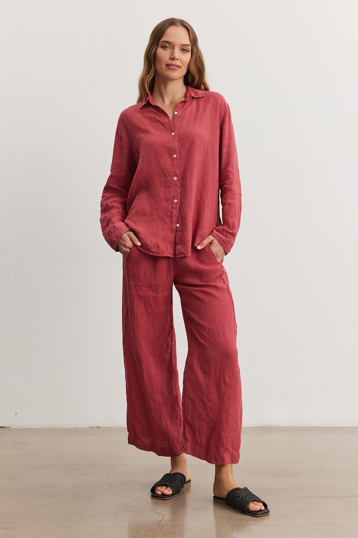 Woman standing in a studio, wearing a red woven linen Velvet by Graham & Spencer Willow Linen Button-Up Shirt and matching pants, paired with black sandals, looking at the camera with a relaxed expression.-36909599883457