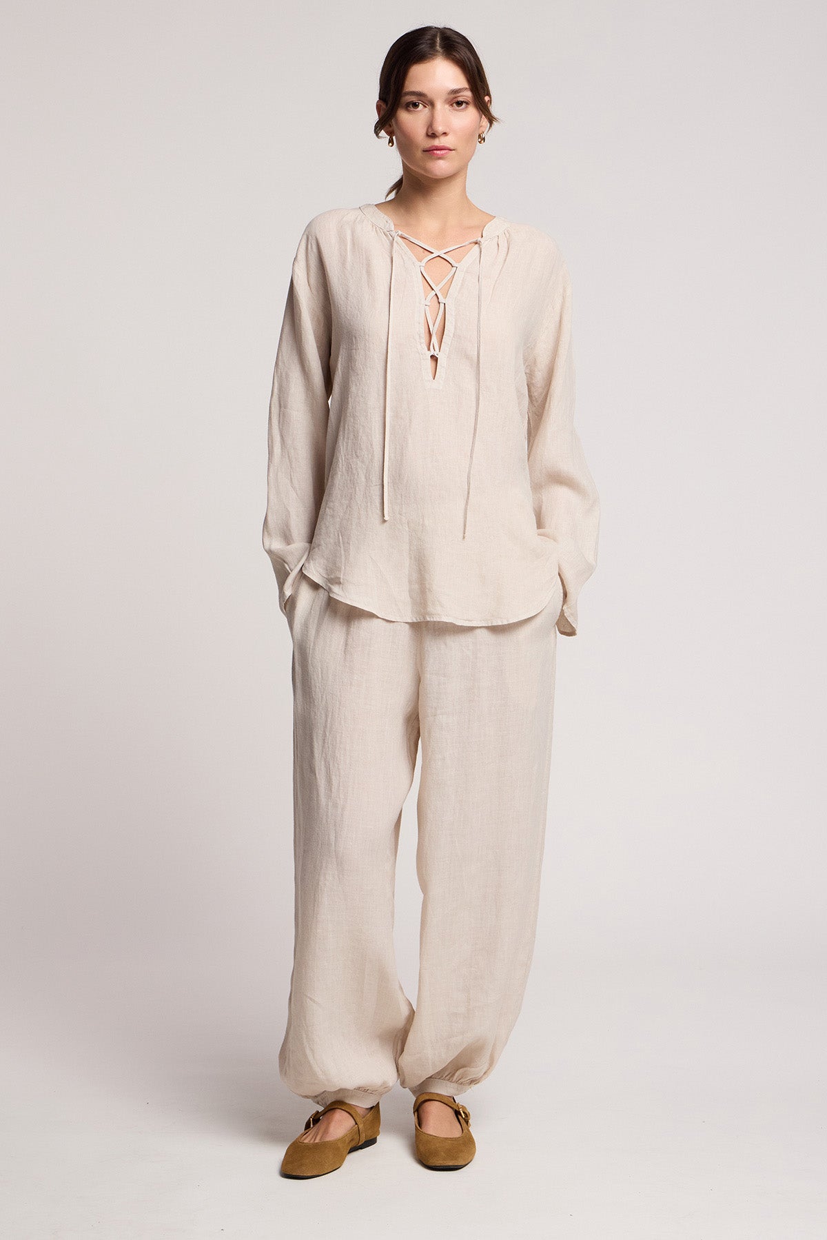 A person stands facing forward wearing a loose, beige linen top with a lace-up neckline and the CAMBER LINEN PANT by Velvet by Graham & Spencer, featuring an elastic waist, paired with brown flat shoes.-41371565064385