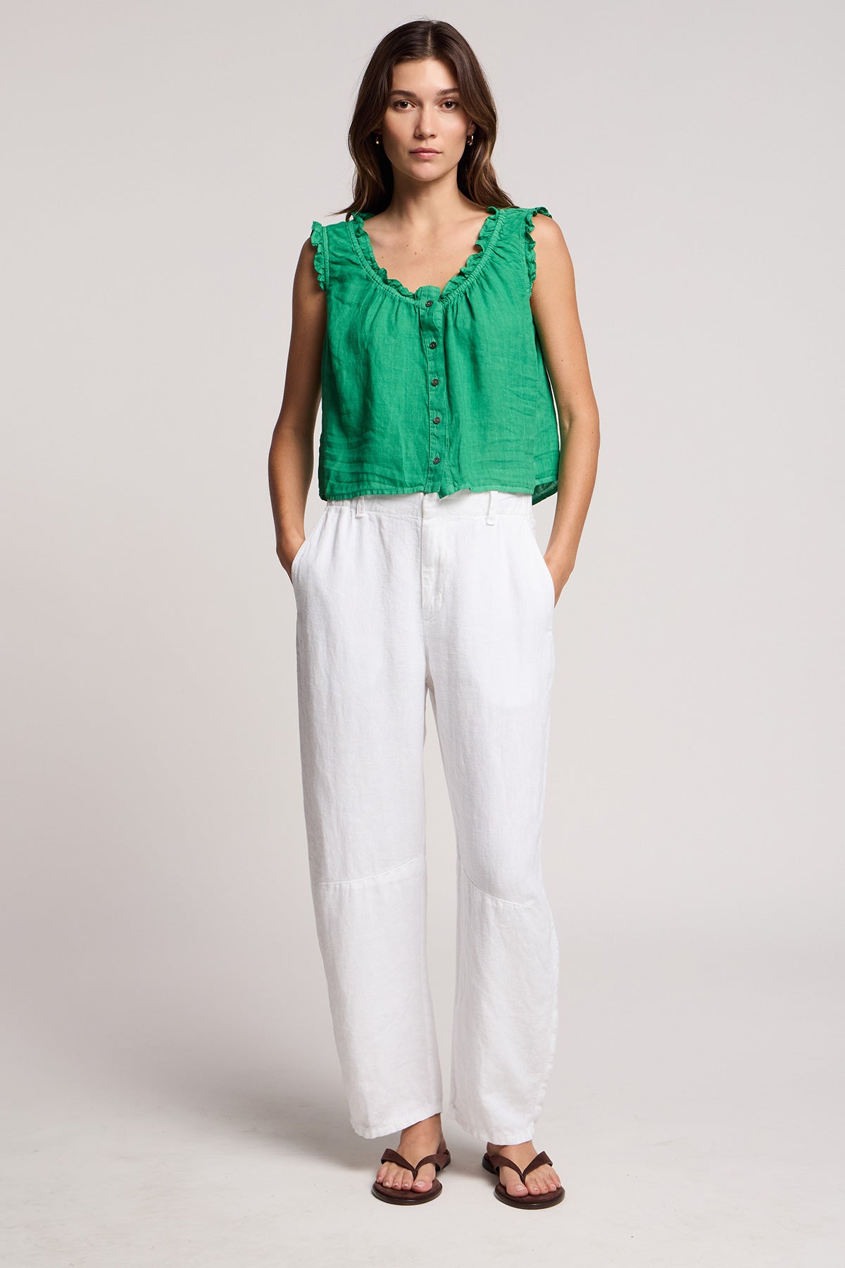 A woman stands with hands in pockets, wearing the TANA LINEN TOP by Velvet by Graham & Spencer—a green sleeveless button-up—paired with white loose pants and brown sandals, against a plain background.-41527238230209
