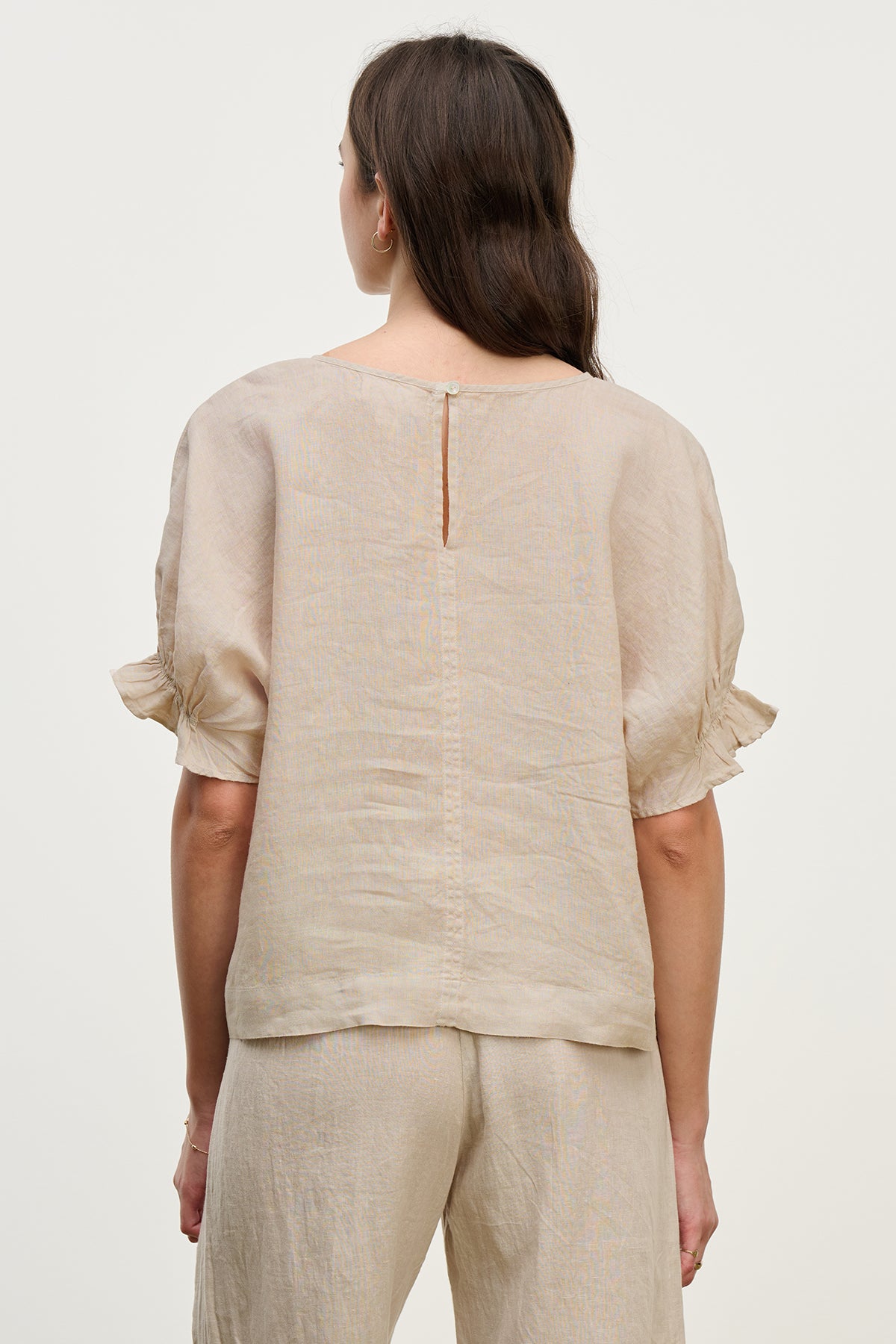 A woman with long brown hair models the EMILY LINEN TOP by Velvet by Graham & Spencer. Her back faces the camera against a light background, and she pairs it with beige pants featuring subtle ruffle cuffs.-38662581747905
