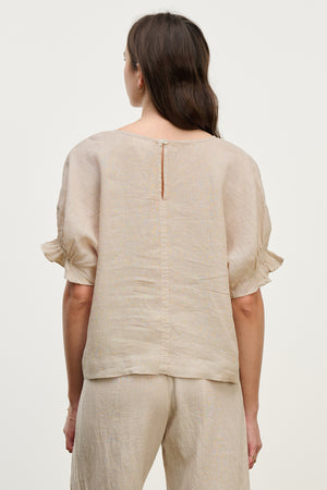 A woman with long brown hair models the EMILY LINEN TOP by Velvet by Graham & Spencer. Her back faces the camera against a light background, and she pairs it with beige pants featuring subtle ruffle cuffs.
