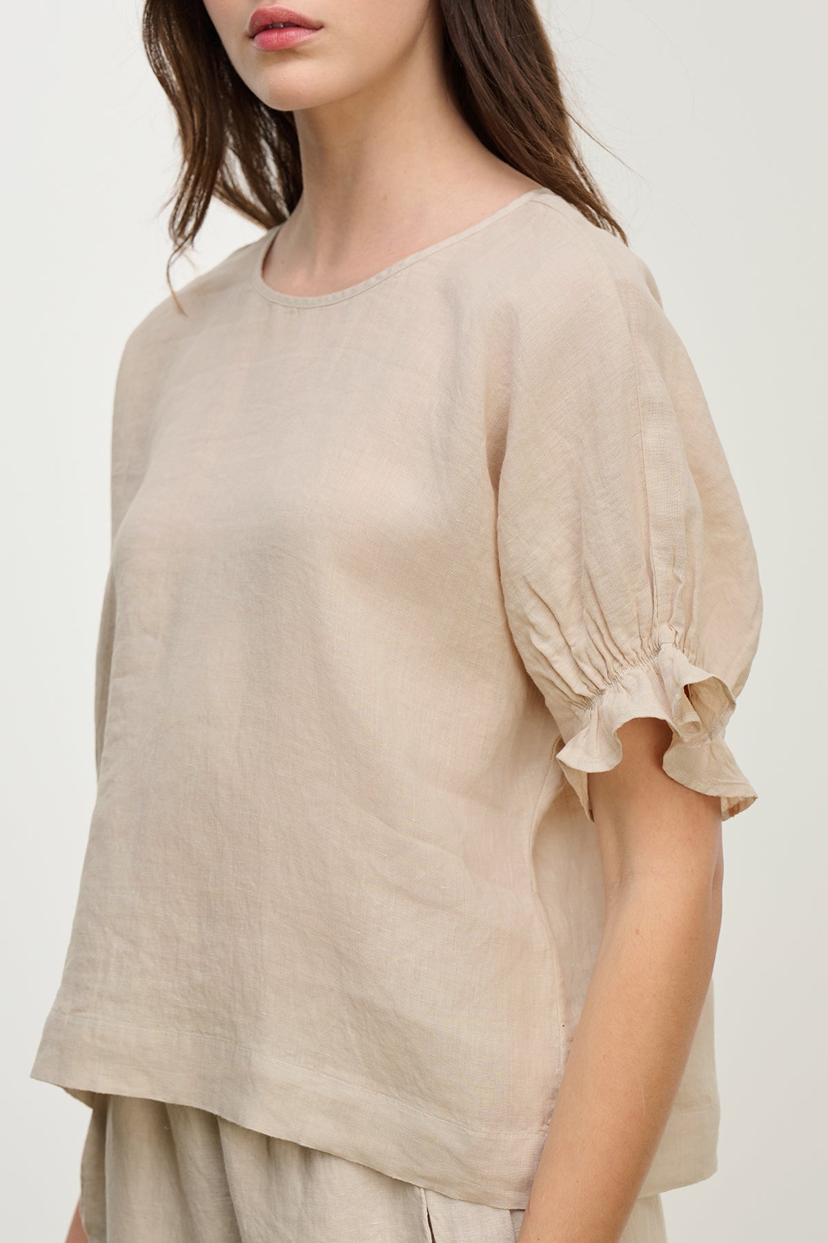 The woman is wearing the Emily Linen Top by Velvet by Graham & Spencer, a beige lightweight top featuring a round neckline and short ruffled sleeves.-38662581780673