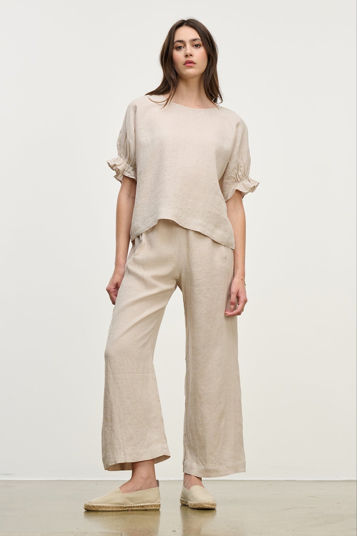 A woman models the Beige LOLA LINEN PANT by Velvet by Graham & Spencer. The outfit features a loose-fitting top and wide-leg, pull-on trousers with an elastic waist, set against a plain white background.