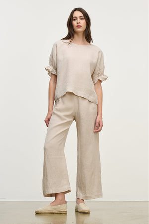A woman models the Beige LOLA LINEN PANT by Velvet by Graham & Spencer. The outfit features a loose-fitting top and wide-leg, pull-on trousers with an elastic waist, set against a plain white background.