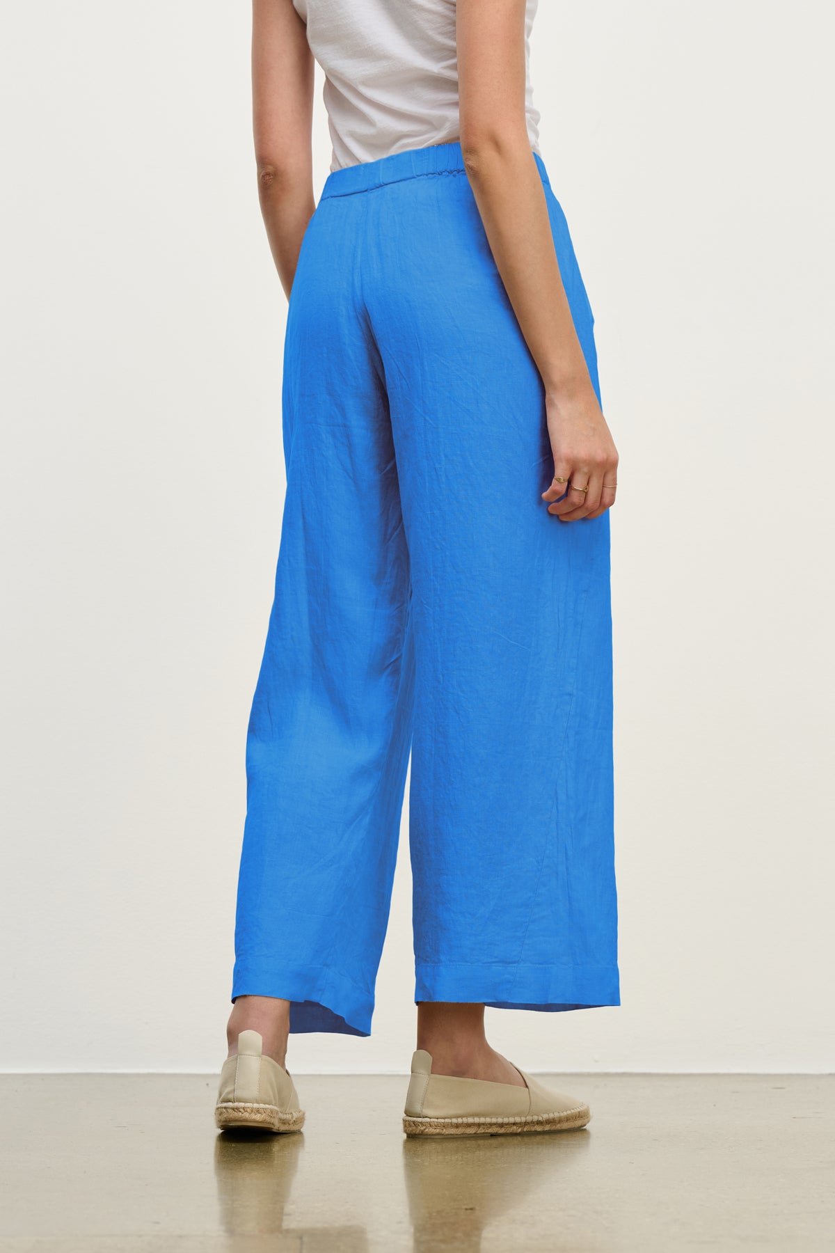 A person stands facing away, wearing the LOLA LINEN PANT by Velvet by Graham & Spencer in bright blue with relaxed legs, paired with a white top and beige espadrilles, set against a plain light background.-39149286621377