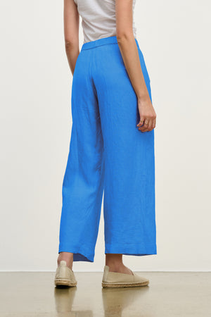 A person stands facing away, wearing the LOLA LINEN PANT by Velvet by Graham & Spencer in bright blue with relaxed legs, paired with a white top and beige espadrilles, set against a plain light background.