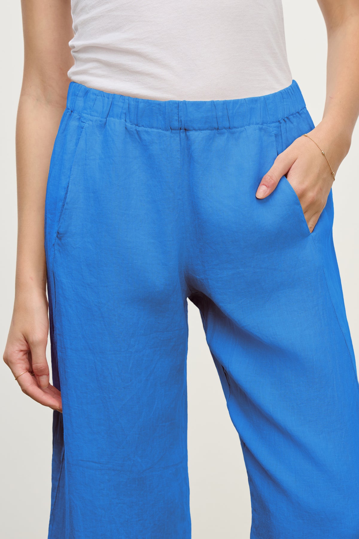 A person wears the LOLA LINEN PANT by Velvet by Graham & Spencer—a bright blue, relaxed leg pant with an elastic waistband—paired with a white top and one hand in the pocket.-39149286654145