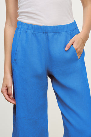A person wears the LOLA LINEN PANT by Velvet by Graham & Spencer—a bright blue, relaxed leg pant with an elastic waistband—paired with a white top and one hand in the pocket.