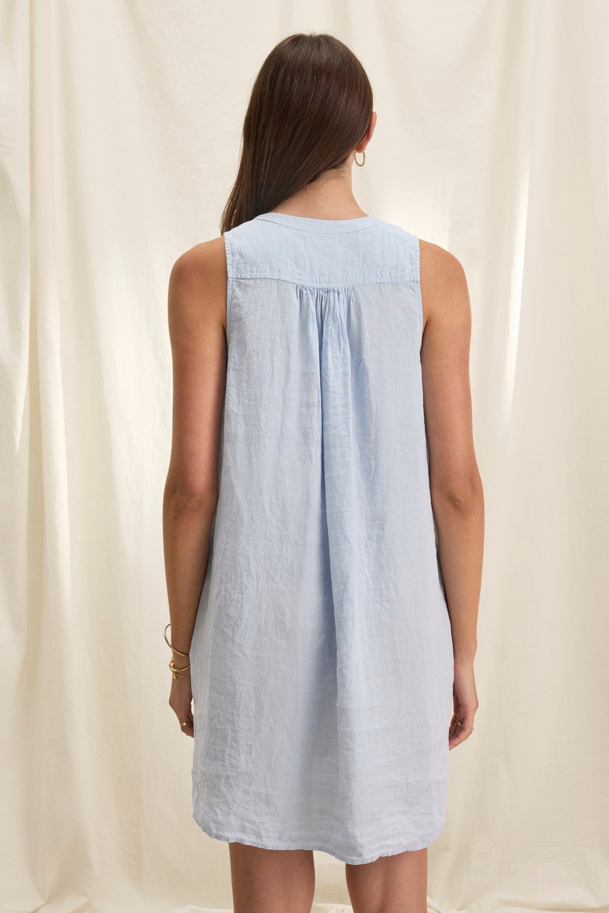 A woman in a sleeveless ADRIANA LINEN DRESS by Velvet by Graham & Spencer, light blue in color, stands with her back to the camera against a light curtain backdrop.