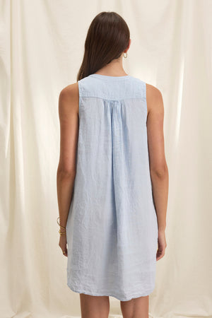 A woman in a sleeveless ADRIANA LINEN DRESS by Velvet by Graham & Spencer, light blue in color, stands with her back to the camera against a light curtain backdrop.