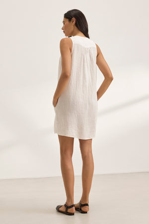 Facing away from a light-colored wall, a person stands with hands in pockets, wearing the ADRIANA LINEN DRESS, a sleeveless woven linen mini with a V-neckline by Velvet by Graham & Spencer.