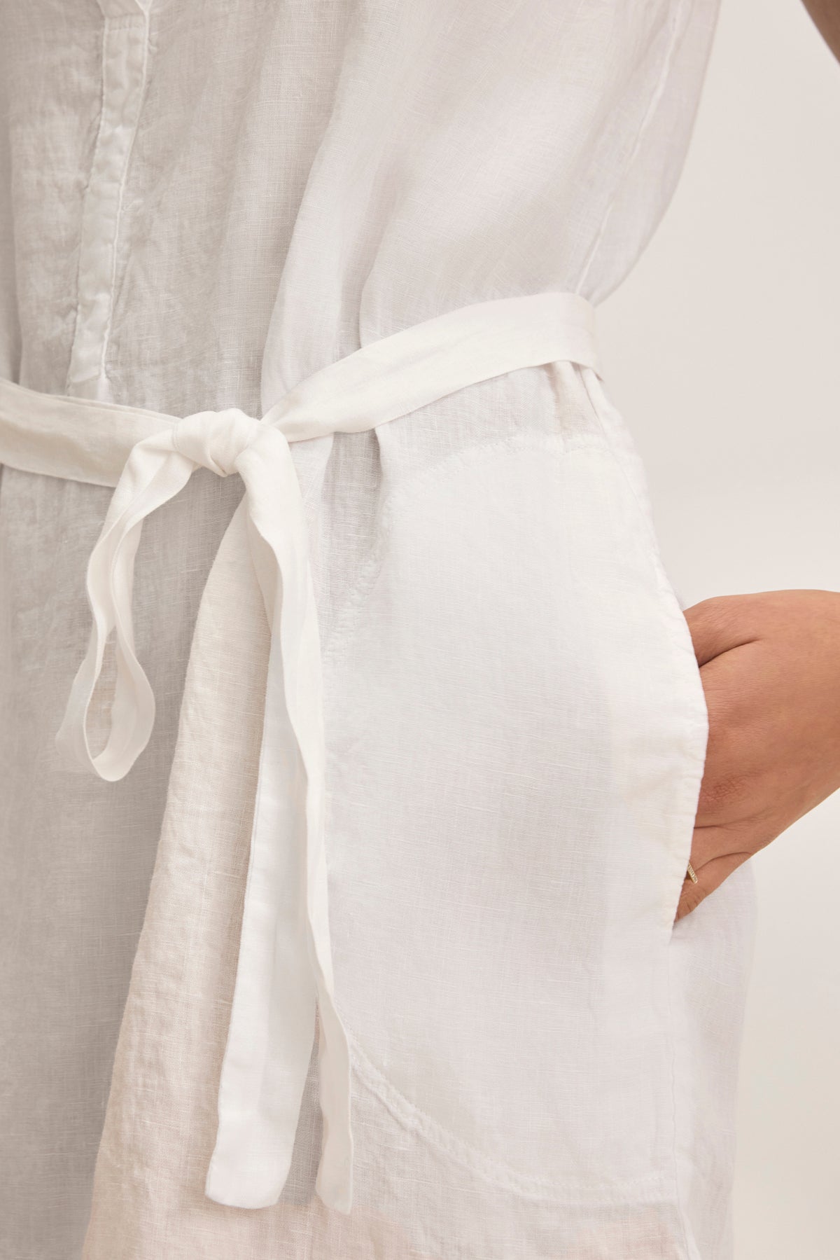 Close-up of a person in the ADRIANA LINEN DRESS by Velvet by Graham & Spencer, showcasing a white woven linen mini dress with a V-neckline, tied waist belt, and right hand in a pocket.