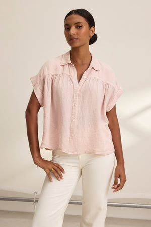 A person stands against a neutral background wearing the ARIA LINEN TOP, a light pink button-up short-sleeve blouse with a relaxed silhouette by Velvet by Graham & Spencer, paired with white pants.