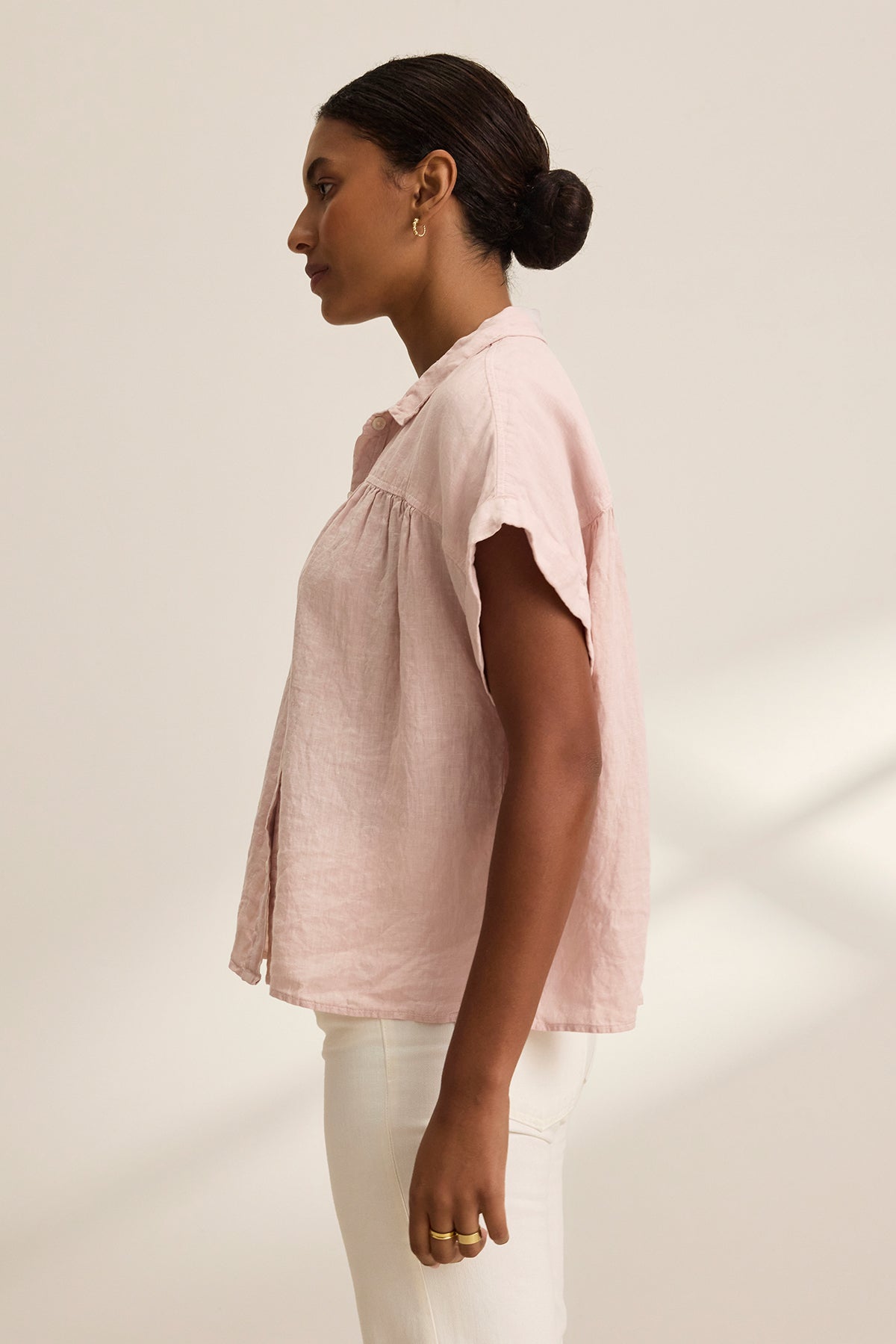 An individual in profile wears the ARIA LINEN TOP by Velvet by Graham & Spencer, a light pink, short-sleeve, button-front shirt featuring a relaxed silhouette, paired with white pants.