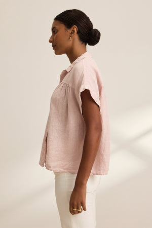 An individual in profile wears the ARIA LINEN TOP by Velvet by Graham & Spencer, a light pink, short-sleeve, button-front shirt featuring a relaxed silhouette, paired with white pants.