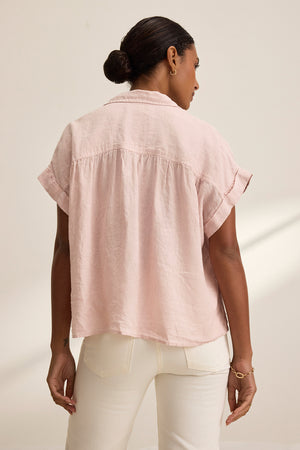 A person with short hair wears the ARIA LINEN TOP by Velvet by Graham & Spencer in light pink, featuring a button front and paired with white pants, exuding a relaxed silhouette while facing away.