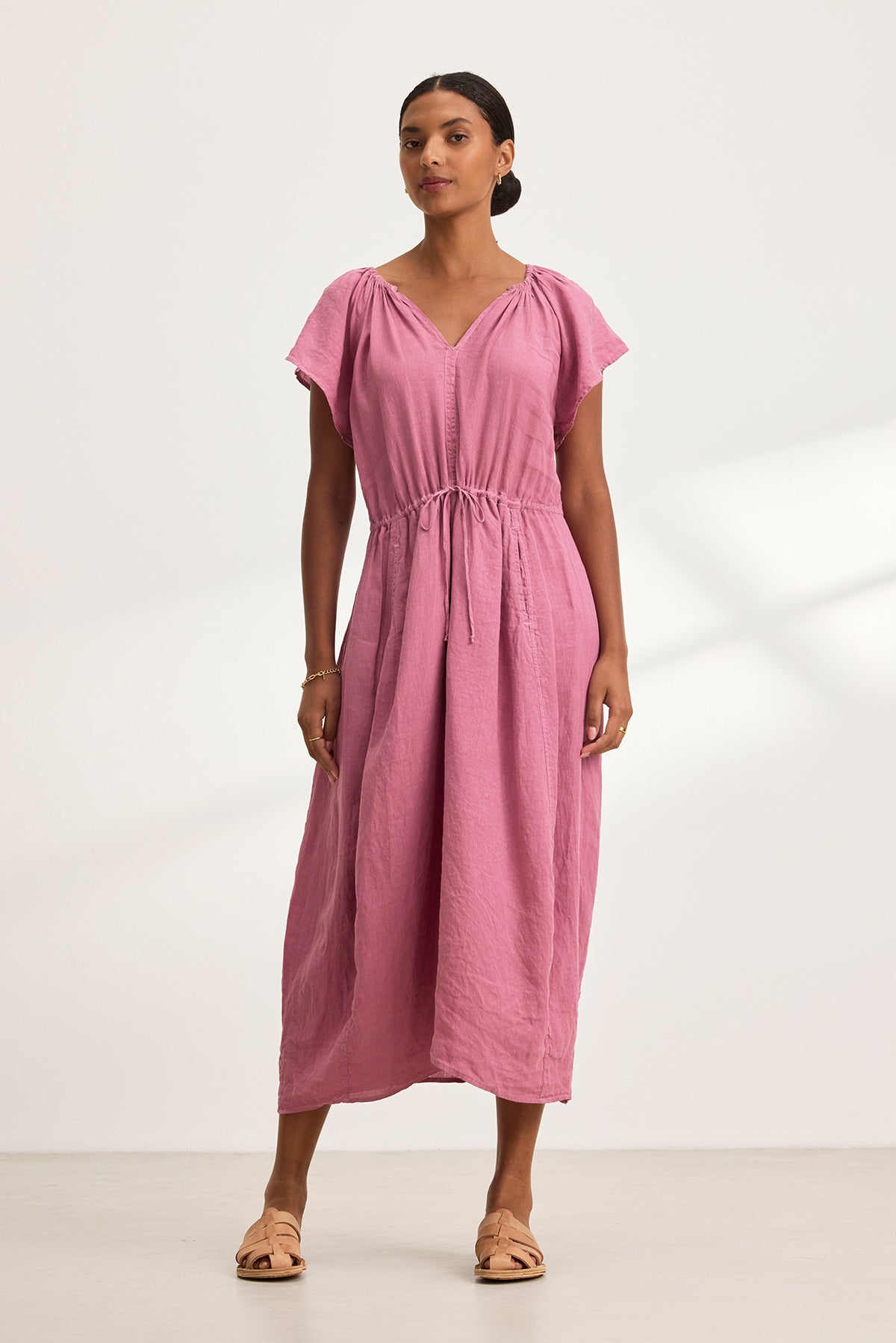 A person wearing the Velvet by Graham & Spencer BALI LINEN DRESS, a pink linen midi piece with flutter sleeves and a drawstring waist, standing on a light-colored floor.