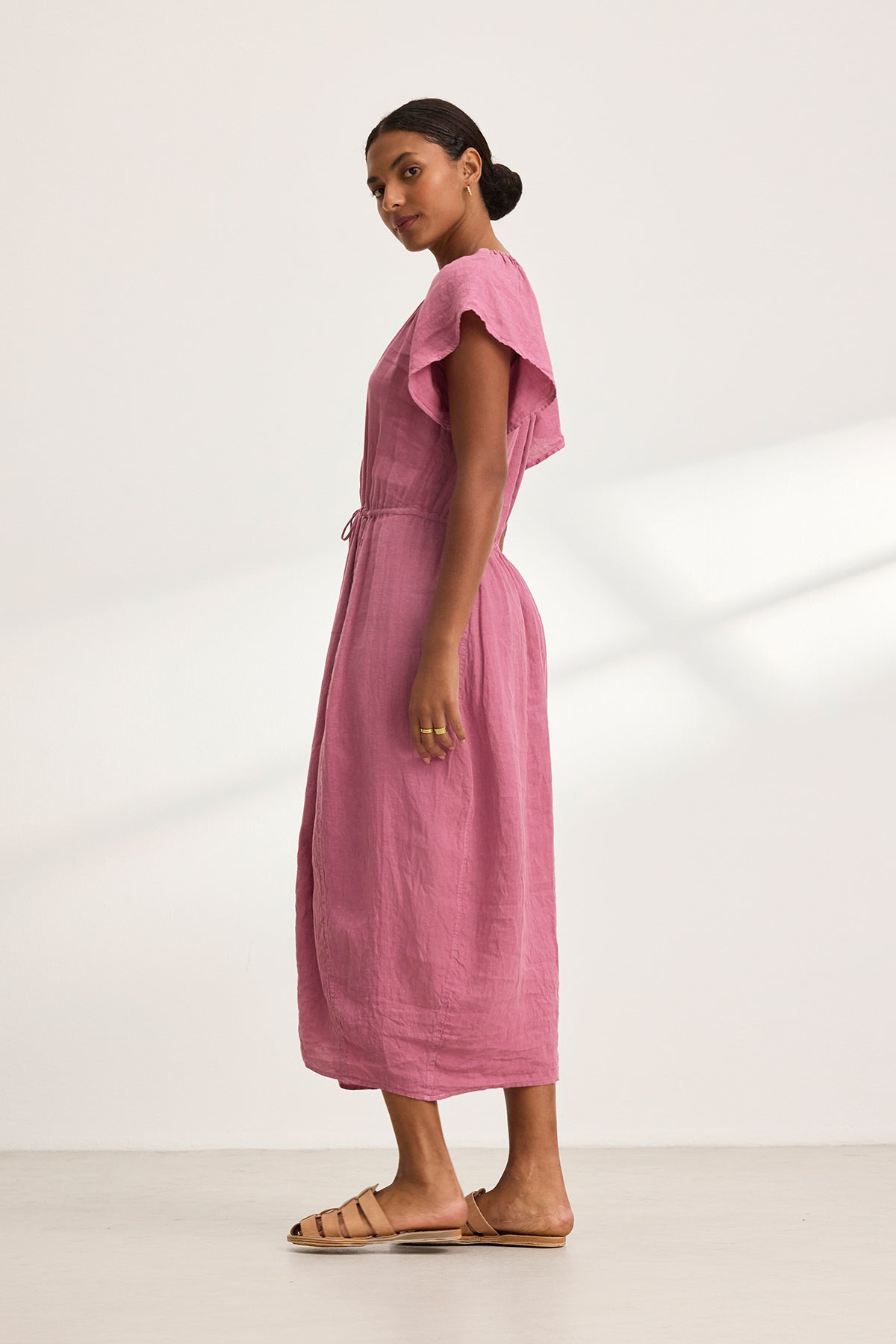 In a brightly lit space, a woman stands sideways, elegantly wearing the BALI LINEN DRESS by Velvet by Graham & Spencer— a pink linen piece with flutter sleeves—paired with brown sandals.-38662638108865