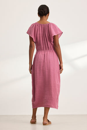 A person with dark hair tied back is seen from behind, wearing the BALI LINEN DRESS by Velvet by Graham & Spencer in pink. The ankle-length dress features flutter sleeves, and they are paired with sandals against a plain white background.