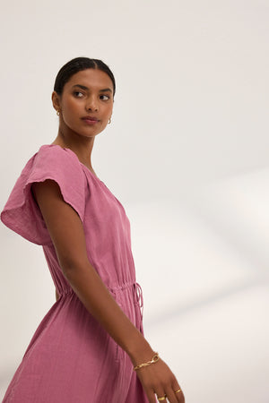 A person in the BALI LINEN DRESS from Velvet by Graham & Spencer, featuring a mauve hue, drawstring waist, and flutter sleeves, gazes to the side against a plain, light background.