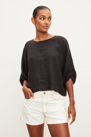 A person wearing the CASS LINEN TOP by Velvet by Graham & Spencer paired with white shorts stands against a plain background.