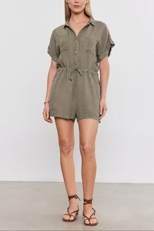A woman in a studio stands wearing the Velvet by Graham & Spencer Clare Linen Romper with rolled-up sleeves and brown strappy sandals. She has blonde hair and a neutral expression.