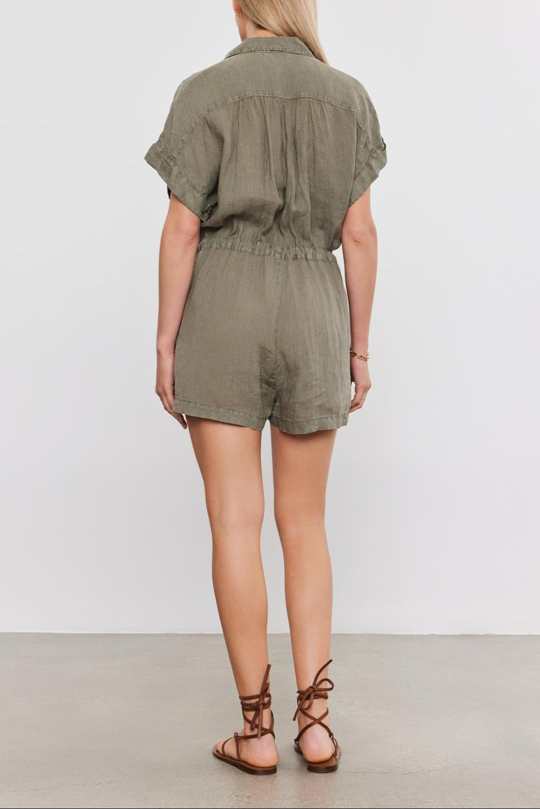 Woman standing facing away, wearing an olive green Velvet by Graham & Spencer CLARE LINEN ROMPER with drawstring waist and brown lace-up sandals.