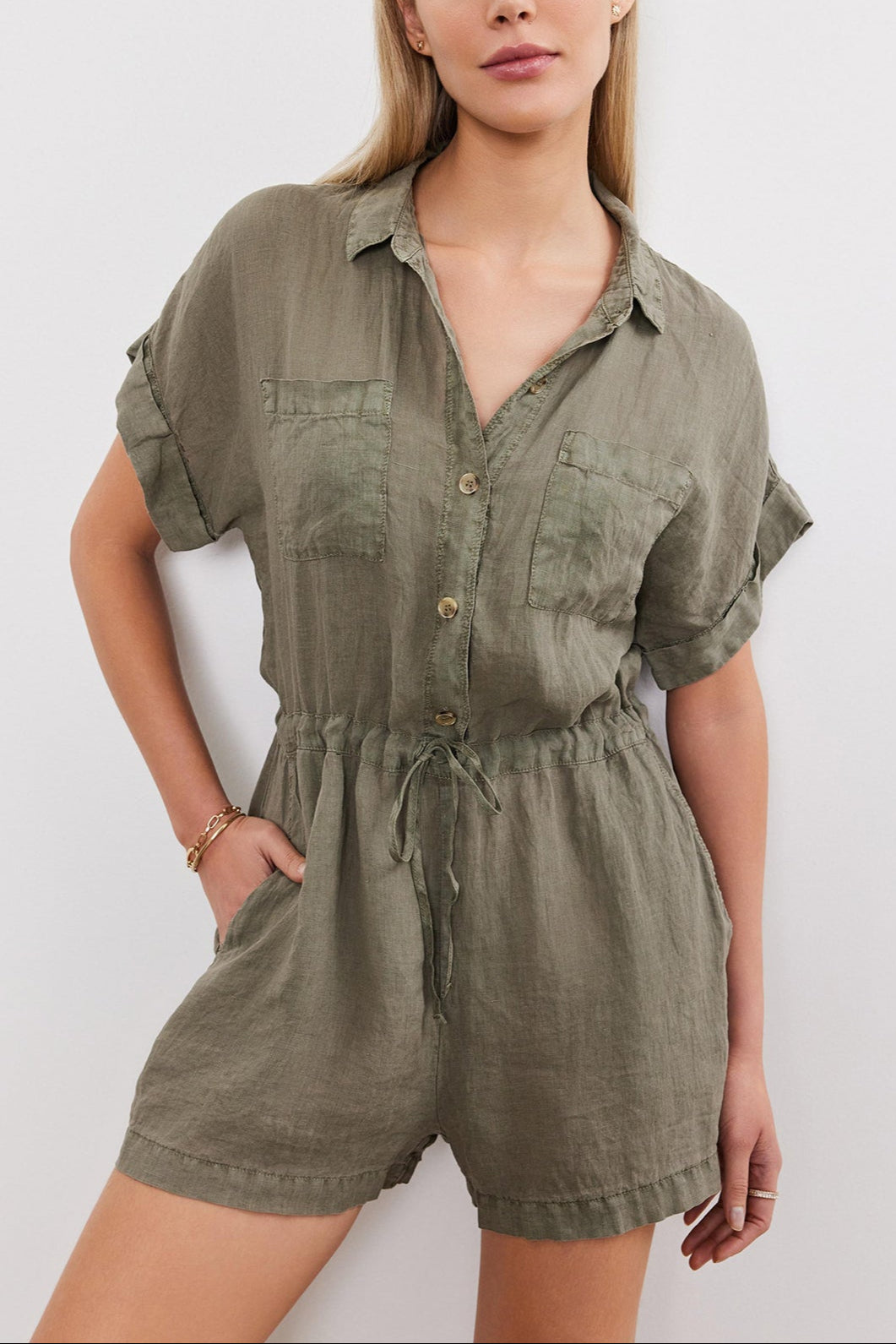 A woman models a short-sleeved, olive green Velvet by Graham & Spencer CLARE LINEN ROMPER with a button-up front and drawstring waist, standing against a plain background.-36910030651585
