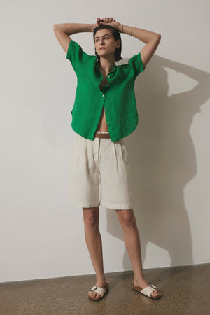 Woman in a green CLAREMONT LINEN SHIRT by Velvet by Jenny Graham and beige shorts posing with hands behind head against a light background.