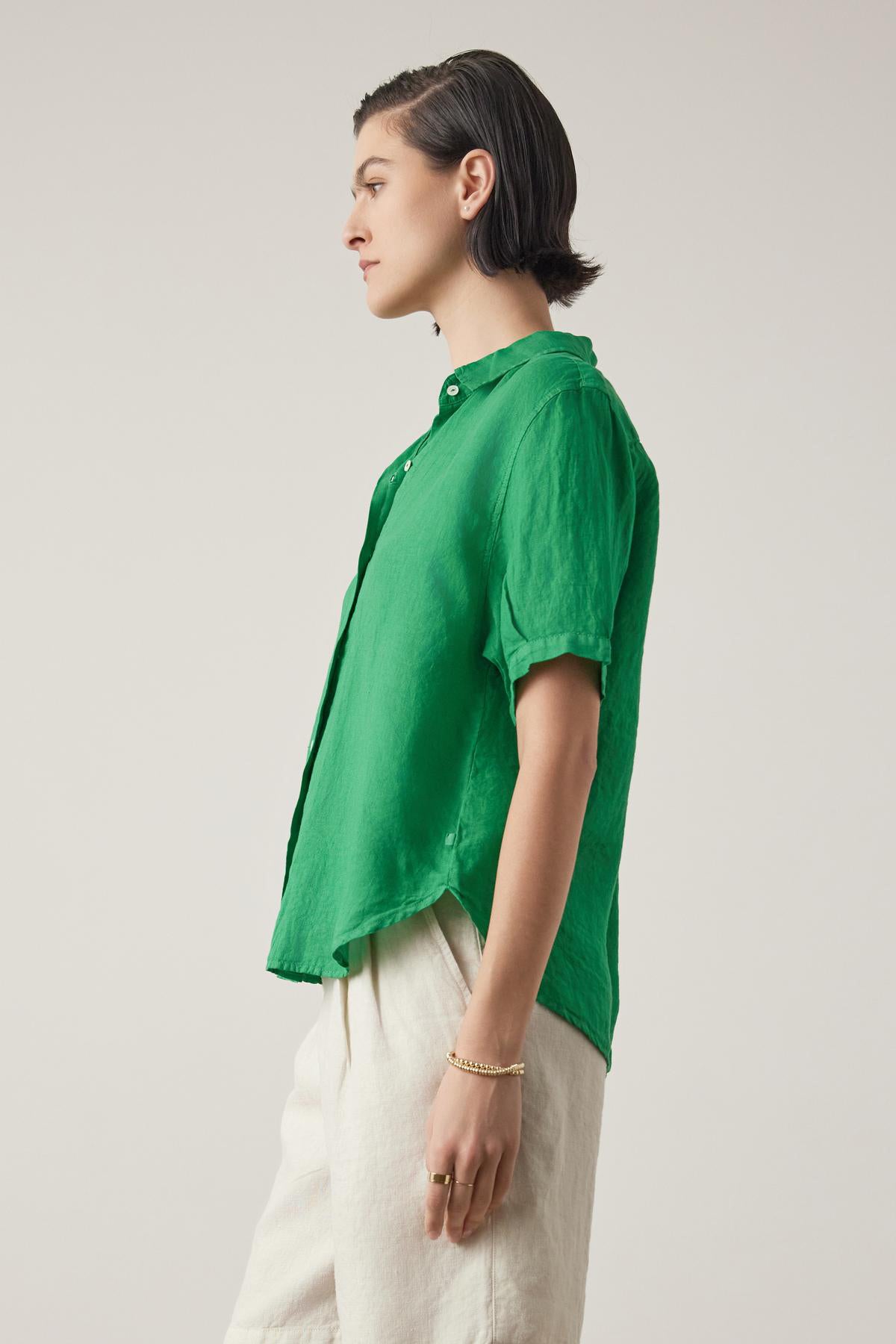   A person wearing a bright green short-sleeved Velvet by Jenny Graham Claremont linen shirt and cream pants, standing in profile against a neutral background. 