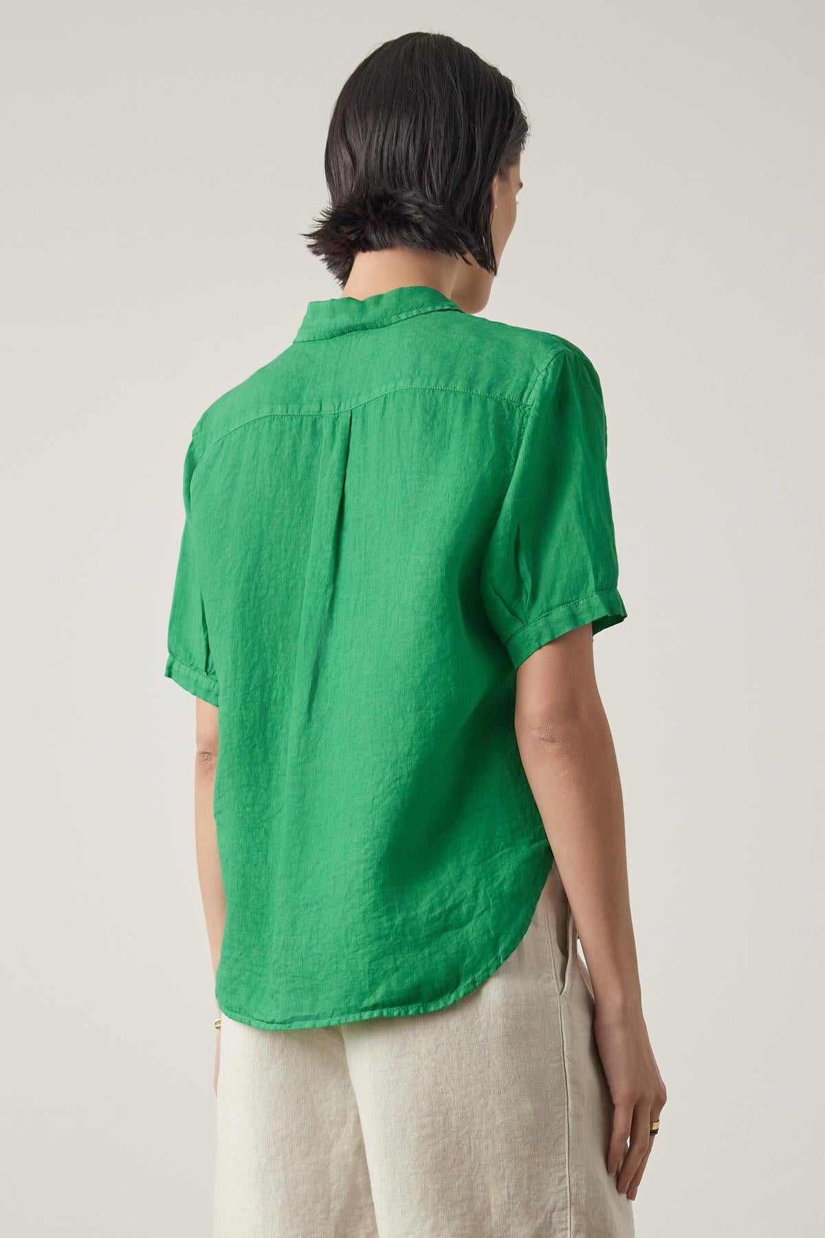   Woman facing away from the camera, wearing a green short-sleeved CLAREMONT LINEN SHIRT by Velvet by Jenny Graham and beige trousers. 