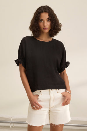 A person with curly hair wears a black EMILY LINEN TOP by Velvet by Graham & Spencer, featuring elastic ruffle cuffs, paired with white shorts in a neutral setting.