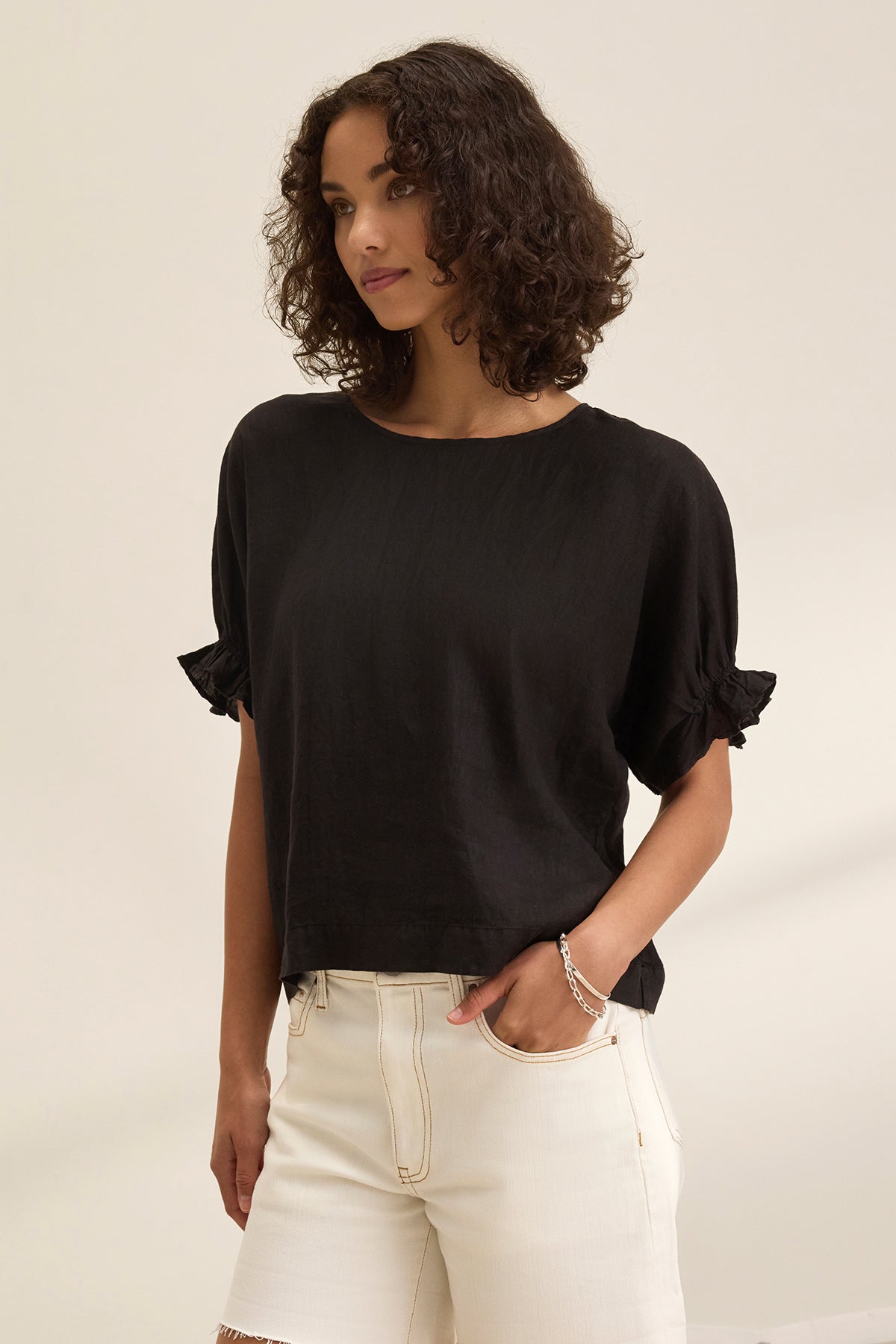A person with curly hair wears the EMILY LINEN TOP by Velvet by Graham & Spencer, featuring elastic ruffle cuffs, paired with white shorts, standing against a plain background.-38662936559809