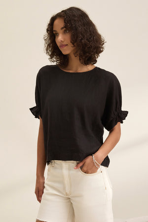 A person with curly hair wears the EMILY LINEN TOP by Velvet by Graham & Spencer, featuring elastic ruffle cuffs, paired with white shorts, standing against a plain background.