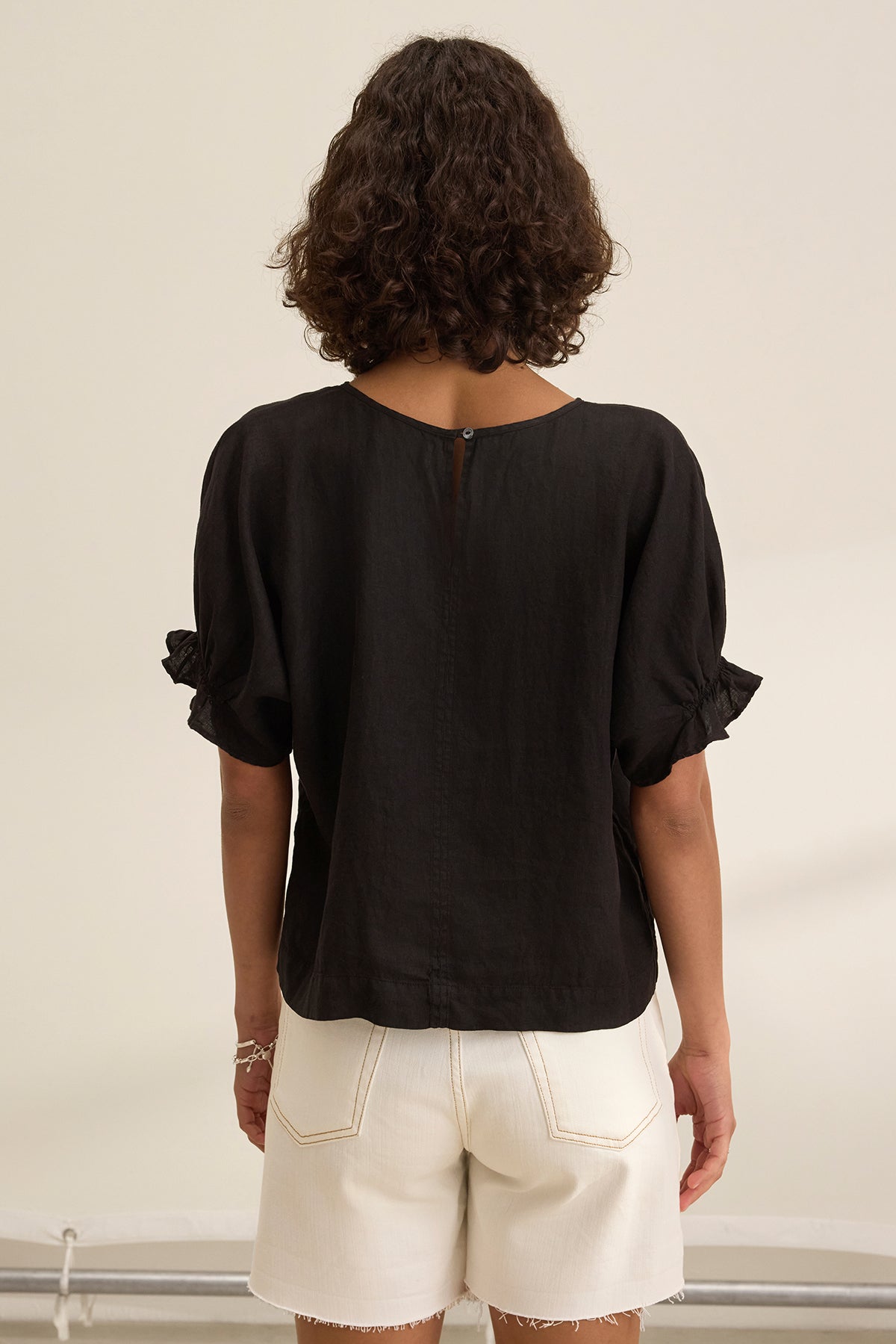 A person with curly hair, seen from the back, wears the EMILY LINEN TOP by Velvet by Graham & Spencer—a black blouse with elastic ruffle cuffs—and white shorts, standing against a neutral background.-38662936592577
