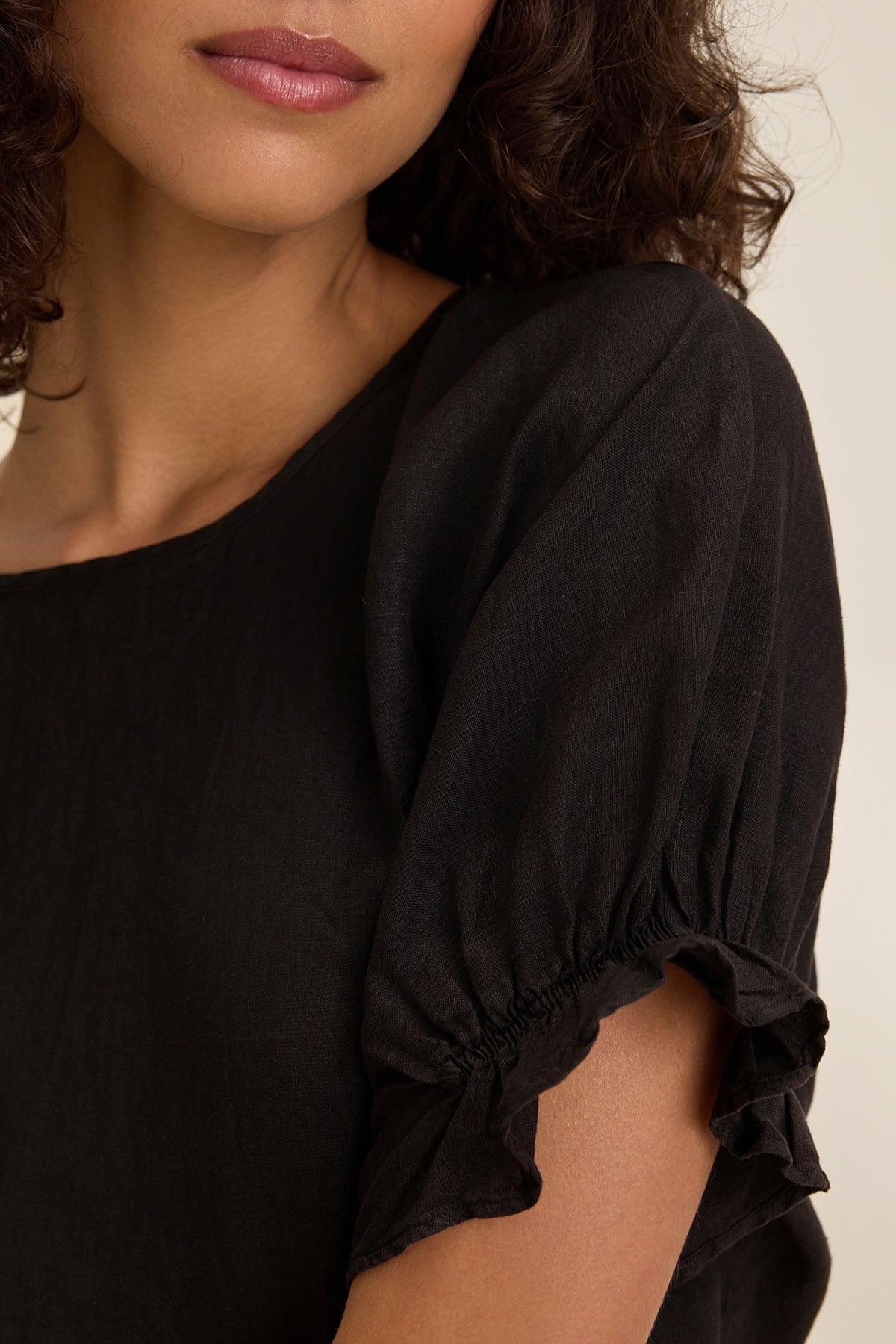 A person with curly hair and neutral lipstick is shown wearing the EMILY LINEN TOP from Velvet by Graham & Spencer. The black, boxy-fit top features gathered short sleeves and elastic ruffle cuffs, visible from neck to upper chest.