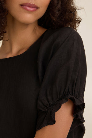A person with curly hair and neutral lipstick is shown wearing the EMILY LINEN TOP from Velvet by Graham & Spencer. The black, boxy-fit top features gathered short sleeves and elastic ruffle cuffs, visible from neck to upper chest.