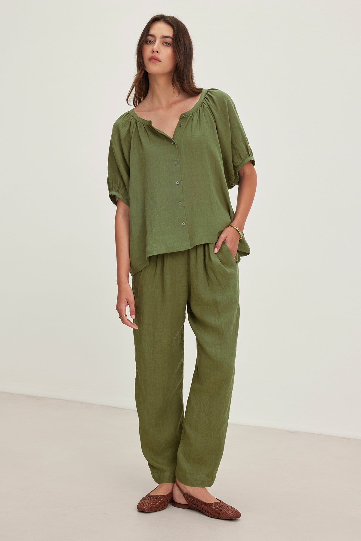 Someone dressed in a loose green blouse and Velvet by Graham & Spencer's GINI LINEN PANT, featuring straight-leg design with an elastic waistband, stands in a minimalist space with neutral flooring and a light background.-38662677364929