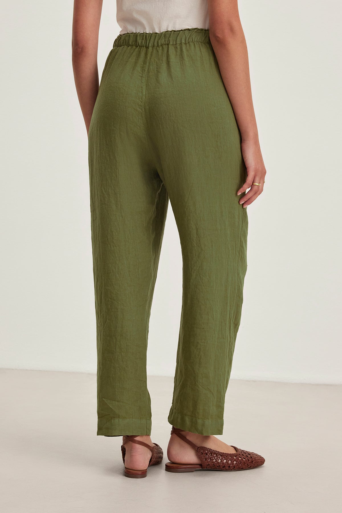 Wearing the GINI LINEN PANT by Velvet by Graham & Spencer, featuring an elastic waistband and straight-leg silhouette in olive green, a person is paired with brown woven sandals and is facing away.