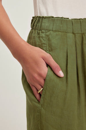 A hand wearing a ring is tucked into the pocket of a GINI LINEN PANT by Velvet by Graham & Spencer, featuring a straight-leg silhouette enhanced by lightweight woven linen.