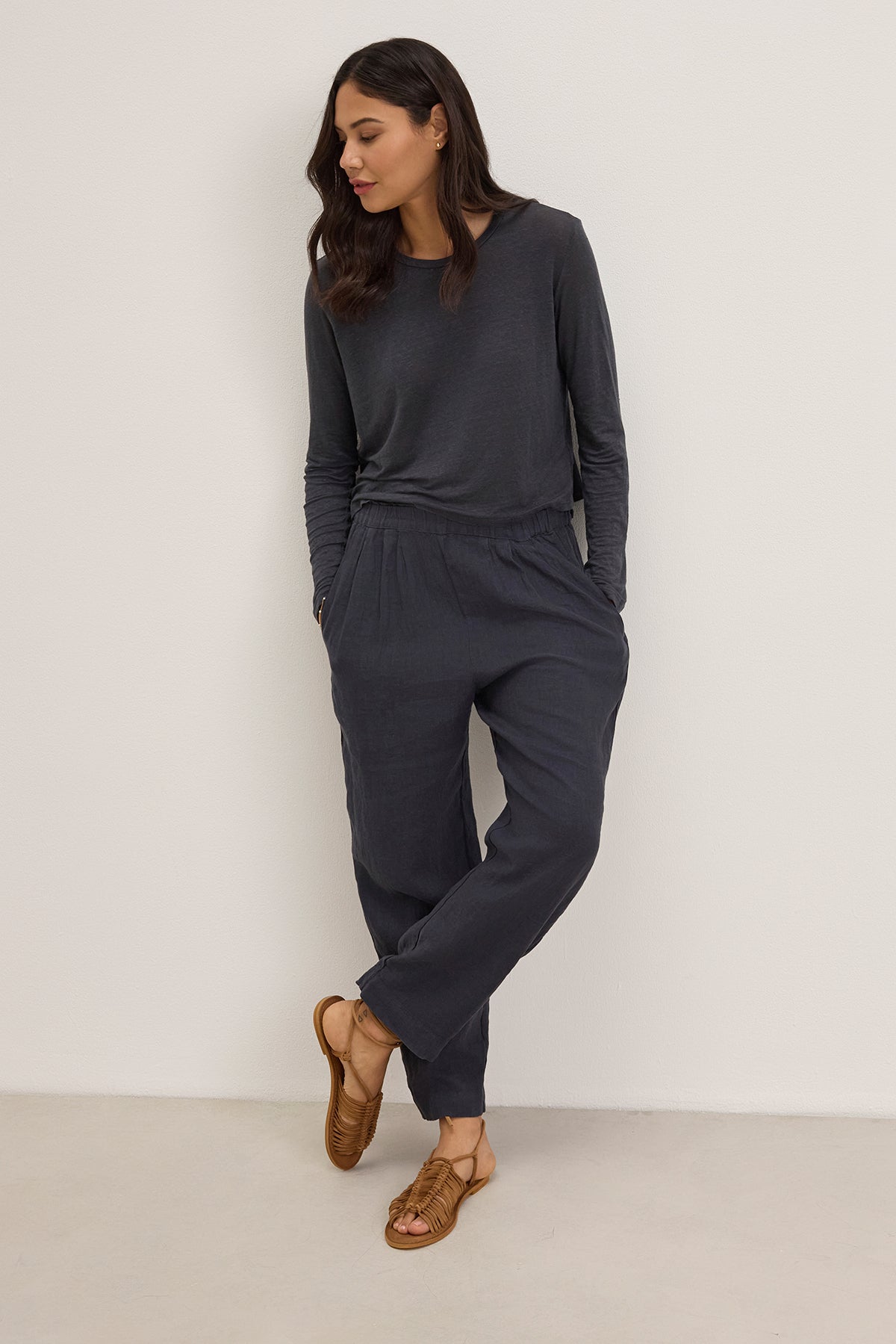 Person in a Velvet by Graham & Spencer dark gray long-sleeved top and GINI LINEN PANT, featuring a straight-leg silhouette with an elastic waistband, leans against a white wall, complemented by brown sandals.