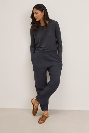 Person in a Velvet by Graham & Spencer dark gray long-sleeved top and GINI LINEN PANT, featuring a straight-leg silhouette with an elastic waistband, leans against a white wall, complemented by brown sandals.